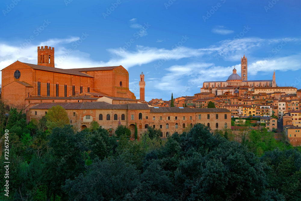 Summer scene of Siena, a medieval town in Tuscany, Italy, with the Dome & Bell Tower of Siena Cathedral in background, landmark Mangia Tower next to Basilica of San Domenico & a forest in foreground
