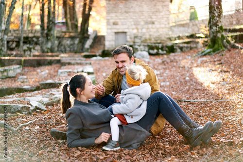 Smiling dad stretches a twig while sitting next to mom with a little girl on her stomach on fallen leaves in the forest