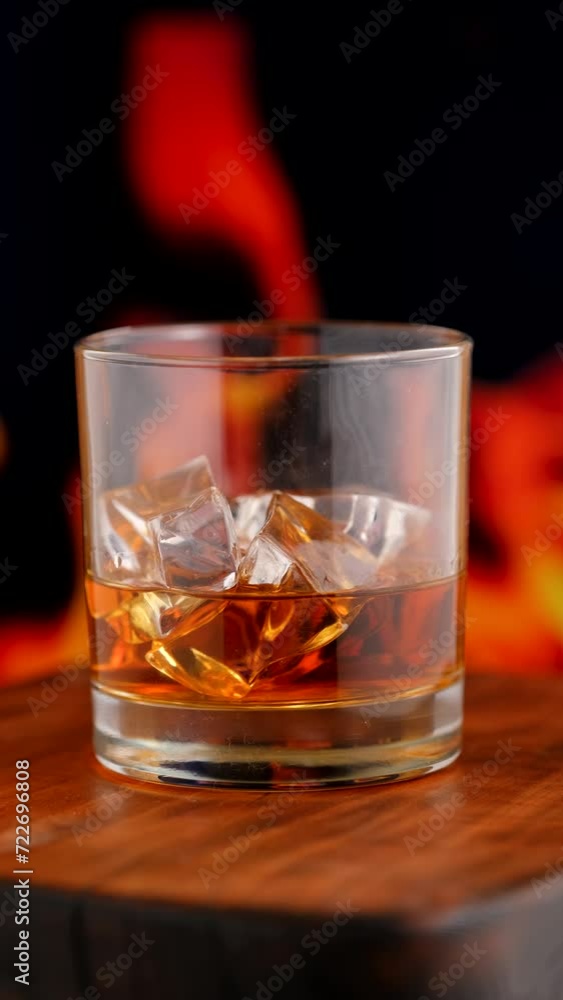 Glass of whisky with ice on wooden tabletop in front of fireplace, closeup shot Glass of whisky with ice on wooden tabletop in front of fireplace, closeup shot