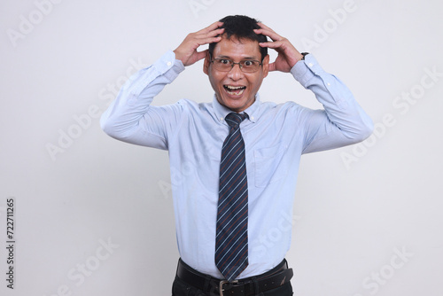 Unhappy Asian man wear glasses, shirt and tie, holding his head and screaming histerically for business failures