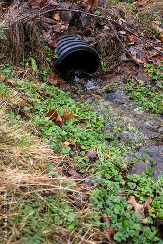 Drainage ditch intake pipe with running water after a heavy rain, surrounded by dead leaves, grass and green weeds 