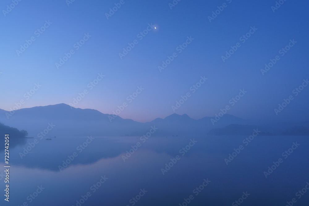 A dawn at Sun Moon Lake in Nantou County, Taiwan. A beautiful morning with half moon over a lake in the mountains. Blue sky with reflection on the lake
