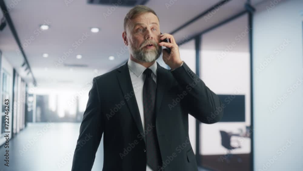 Confident Middle Aged Businessman in a Suit Walking in a Modern Office Corridor, Talking on a Phone Call with Corporate Business Partners. Successful Company CEO Planning Profitable Work Projects Confident Middle Aged Businessman in a Suit Walking in a Modern Office Corridor, Talking on a Phone Call with Corporate Business Partners. Successful Company CEO Planning Profitable Work Projects