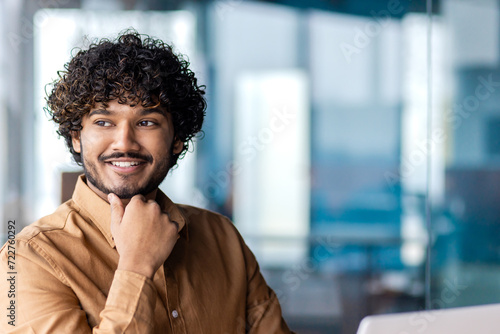 Portrait of muslim male in mustard shirt looking aside and resting head on hand with clenched fist. Smiling man daydreaming and imagining upcoming vacations during working day at modern office.