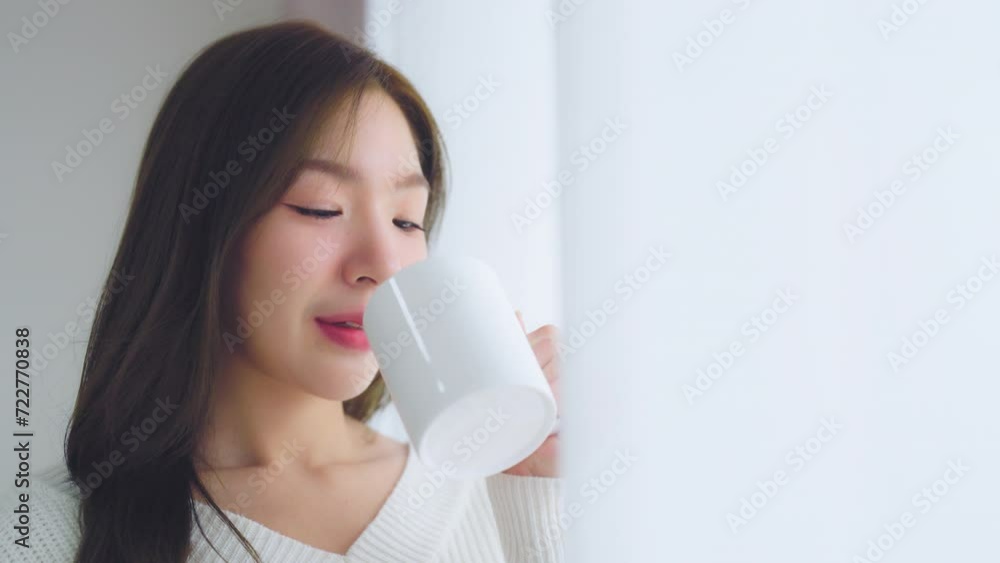 Young asian woman standing beside window and holding mug in bedroom at home, She drinking milk after wake up in the morning Young asian woman standing beside window and holding mug in bedroom at home, She drinking milk after wake up in the morning