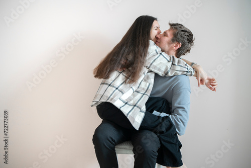 A playful young couple shares an intimate hug while balanced on a chair, their expressions reflecting happiness and affectionate enjoyment.