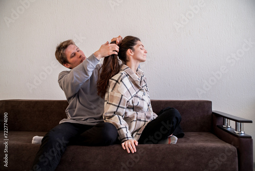 A man focuses intently as he styles a womans long hair while they both sit comfortably on a sofa in a home environment.