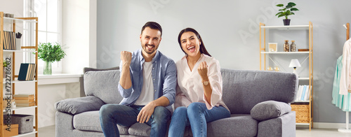 Excited couple celebrating success while sitting on sofa at home. Overjoyed happy young man and woman in casual wear raising clenched fists in excitement doing winner gesture photo