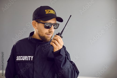 Security guard man in black uniform cap, jacket and sunglasses standing inside house and talking on modern portable wireless two way walkie talkie transceiver radio set device. Property safety concept photo