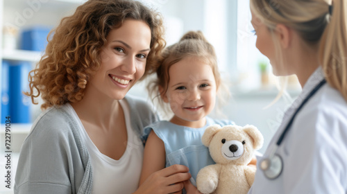 young girl with curly hair holding a teddy bear, accompanied by a female healthcare professional