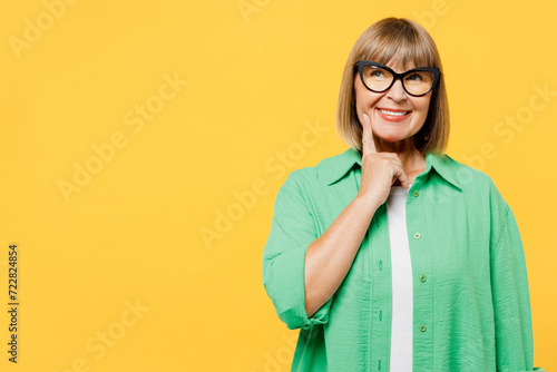 Elderly fun blonde woman 50s years old wear green shirt glasses casual clothes put hand prop up on chin, lost in thought and conjectures isolated on plain yellow background studio. Lifestyle concept.