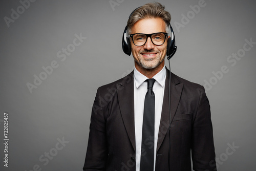 Happy employee operator business man wears black suit set microphone headset for helpline assistance sit work at call center office desk with pc computer isolated on grey background studio portrait.