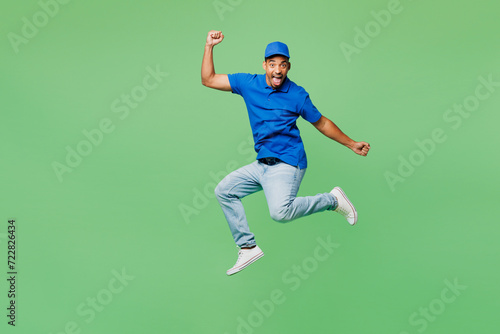 Full body surprised shocked fun delivery guy employee man wear blue cap t-shirt uniform workwear work as dealer courier jump high do winner gesture isolated on plain green background. Service concept.