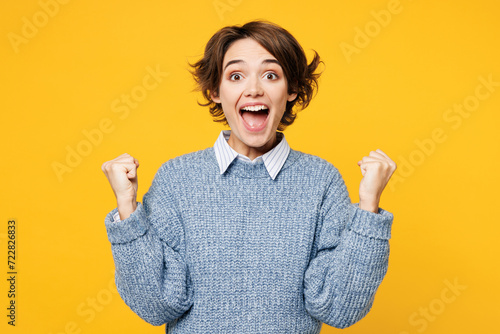 Young overjoyed happy woman wears grey knitted sweater shirt casual clothes doing winner gesture celebrate clench fists say yes isolated on plain yellow background studio portrait. Lifestyle concept. photo