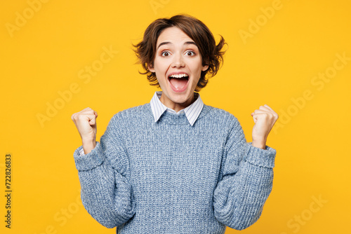 Tableau sur toile Young overjoyed happy woman wears grey knitted sweater shirt casual clothes doing winner gesture celebrate clench fists say yes isolated on plain yellow background studio portrait
