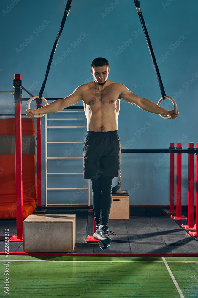 Full-length image of young shirtless man training in gym, doing exercises with gymnastic rings. Strong relief body. Concept of active and healthy lifestyle, body care, fitness, sport