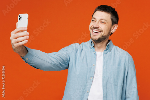 Young man wears blue shirt white t-shirt casual clothes doing selfie shot on mobile cell phone post photo on social network isolated on plain red orange background studio portrait. Lifestyle concept.