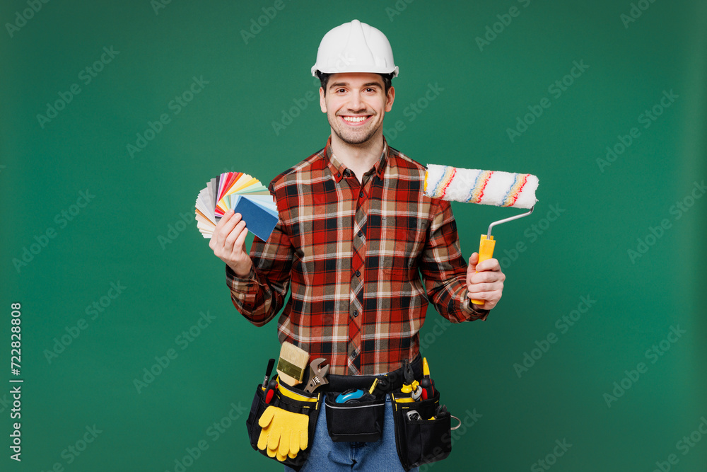 Young laborer man he wear red shirt hardhat hat hold color wallpaper palette paint brush isolated on plain green background. Instruments accessories for renovation apartment room. Repair home concept. Young laborer man he wear red shirt hardhat hat hold color wallpaper palette paint brush isolated on plain green background. Instruments accessories for renovation apartment room. Repair home concept.