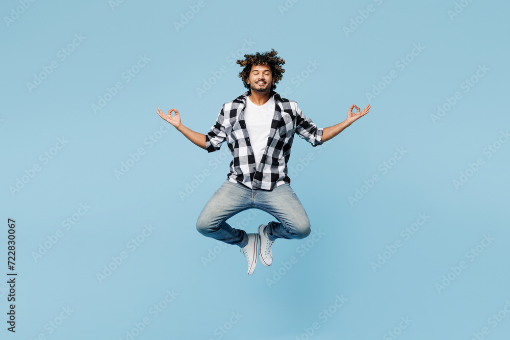 Full body young Indian man wear shirt white t-shirt casual clothes jump high hold spread hands in yoga om aum gesture relax meditate try calm down isolated on plain blue background. Lifestyle concept Full body young Indian man wear shirt white t-shirt casual clothes jump high hold spread hands in yoga om aum gesture relax meditate try calm down isolated on plain blue background. Lifestyle concept