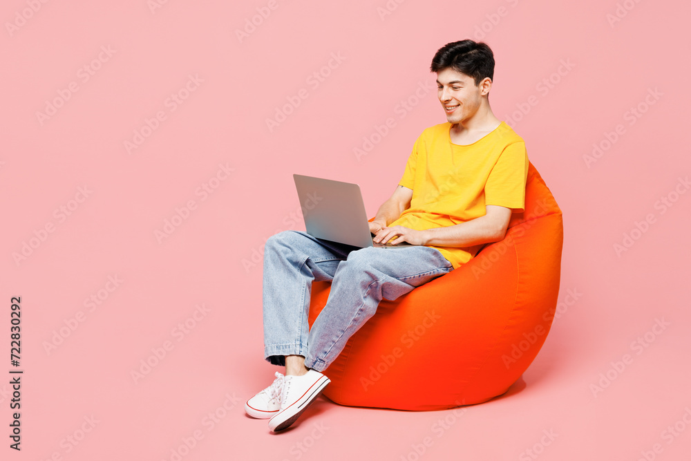 Full body young IT man he wearing yellow t-shirt casual clothes sit in bag chair hold use work on laptop pc computer isolated on plain pastel light pink background studio portrait. Lifestyle concept. Full body young IT man he wearing yellow t-shirt casual clothes sit in bag chair hold use work on laptop pc computer isolated on plain pastel light pink background studio portrait. Lifestyle concept.