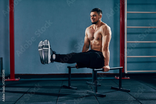 Full-body workout. Muscular, shirtless athletic man training in modern gym, doing L-sit exercise on parallel bars. Concept of active and healthy lifestyle, body care, fitness, sport