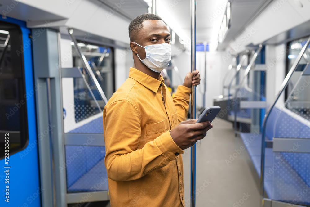 An African-American businessman in a protective mask with a mobile phone rides in a subway car