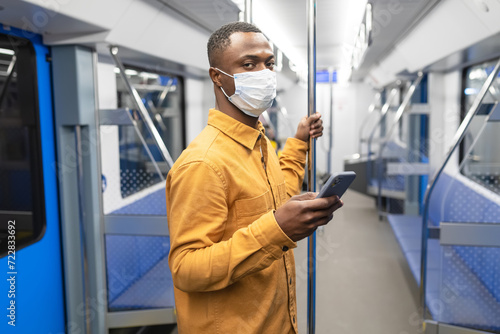 An African-American businessman in a protective mask with a mobile phone rides in a subway car