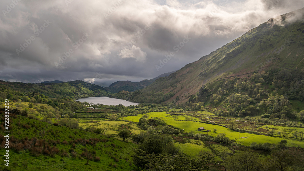 Moody clouds roll over a lush, green valley, surounded by the mountains of Snowdonia, north Wales, UK, with the sun highlighting parts of the vally floor