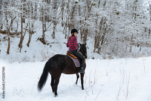 A beautiful brunette girl in a plaid black-and-white shirt walks with a big black horse in a snowy park. A woman in a helmet for riding walks in a snowy forest with a thoroughbred horse