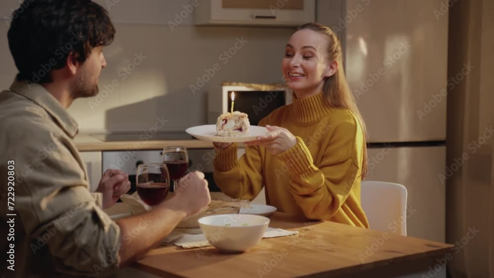 A delighted woman expresses surprise and happiness as she receives a slice of birthday cake with a lit candle from a man in a warm, homely kitchen environment. A delighted woman expresses surprise and happiness as she receives a slice of birthday cake with a lit candle from a man in a warm, homely kitchen environment.