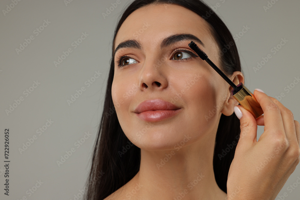 Beautiful young woman applying mascara on grey background, closeup Beautiful young woman applying mascara on grey background, closeup