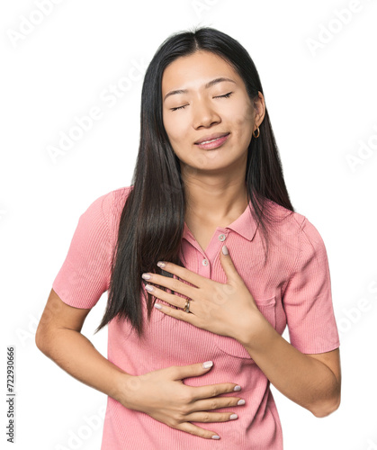 Young Chinese woman in studio setting laughs happily and has fun keeping hands on stomach.