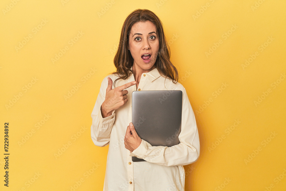 Middle-aged woman with laptop on yellow pointing to the side Middle-aged woman with laptop on yellow pointing to the side