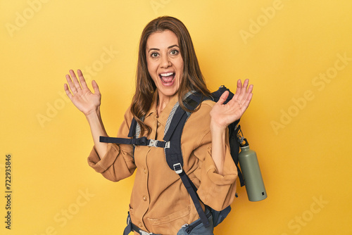 Middle aged woman prepped for hiking, yellow studio shot receiving a pleasant surprise, excited and raising hands.