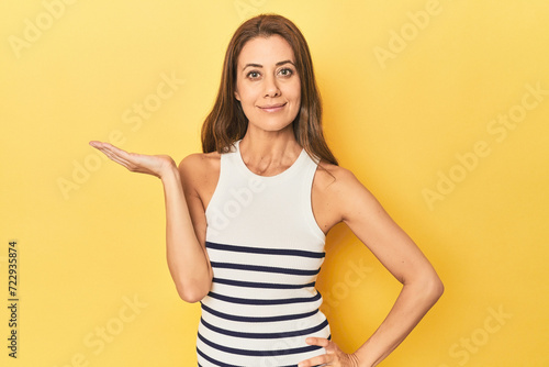 Middle-aged caucasian woman on yellow showing a copy space on a palm and holding another hand on waist.