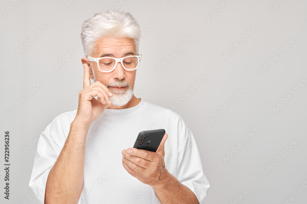 Portrait of gray-haired man in white T-shirt looks on mobile phone and thinks. Person with smartphone isolated on white background. Portrait of gray-haired man in white T-shirt looks on mobile phone and thinks. Person with smartphone isolated on white background.