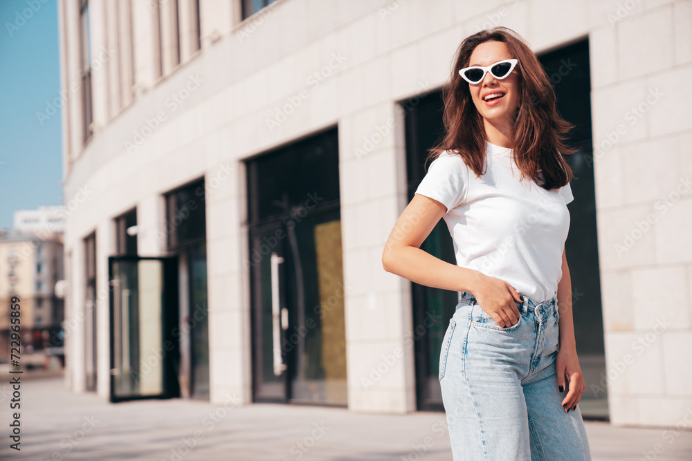 Portrait of young beautiful smiling hipster woman in trendy summer white t-shirt and jeans clothes. Sexy carefree model posing on the street background at sunset. Positive model outdoors in sunglasses