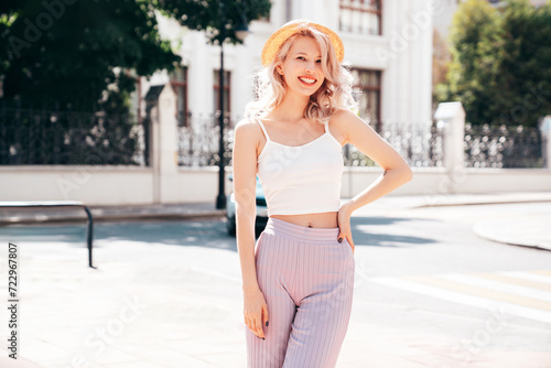 Young beautiful smiling blond hipster woman in trendy summer clothes. Carefree female posing in the street at sunny day. Positive model outdoors at sunset. Cheerful and happy in hat