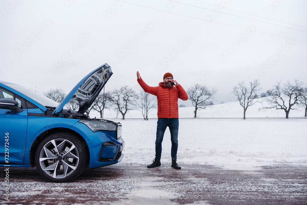 Angry man standing by electric car, battery run out of power before reaching destination. Man phone calling for help looking undre the car hood. Angry man standing by electric car, battery run out of power before reaching destination. Man phone calling for help looking undre the car hood.