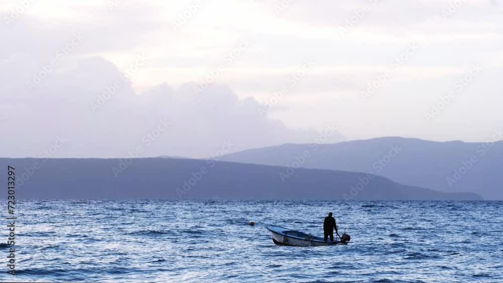 Unrecognizable fisherman in a boat sailing on the sea. Early morning fishing near the coast. People at work concept. Cloudy sky, pastel colours, calm ocean scenery with mountains in the background. Unrecognizable fisherman in a boat sailing on the sea. Early morning fishing near the coast. People at work concept. Cloudy sky, pastel colours, calm ocean scenery with mountains in the background.
