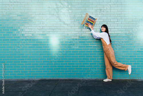 Smiling young woman holding abacus in front of turquoise brick wall photo