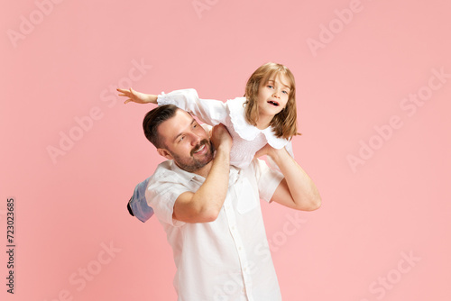 Man with playful smile carrying young girl on his shoulders, both simulating flight against pink pastel background. Concept of International Day of Happiness, childhood and parenthood. Ad photo