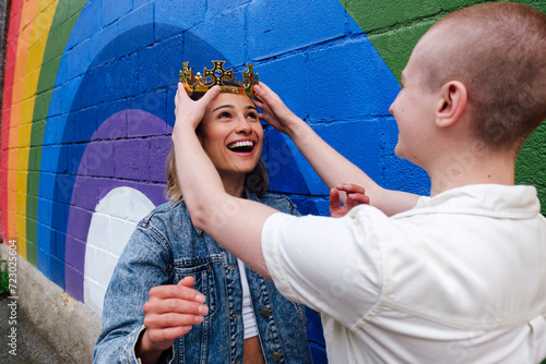 Non-binary person adjusting crown on lesbian woman's head by rainbow wall photo