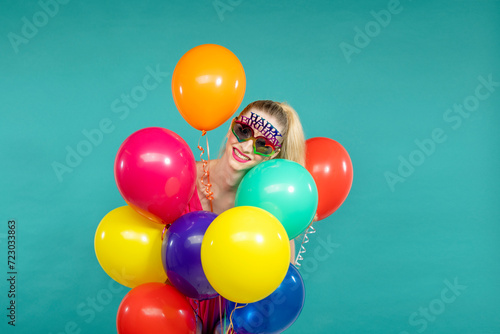 Smiling young woman wearing birthday eyeglasses holding balloons against green background photo