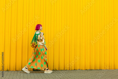 Senior woman carrying fruits in mesh bag and walking near yellow wall photo
