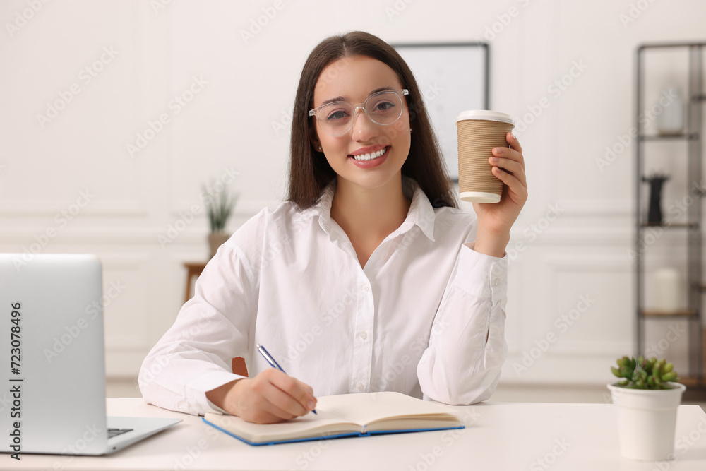 Young woman with cup of coffee writing in notebook at white table indoors Young woman with cup of coffee writing in notebook at white table indoors