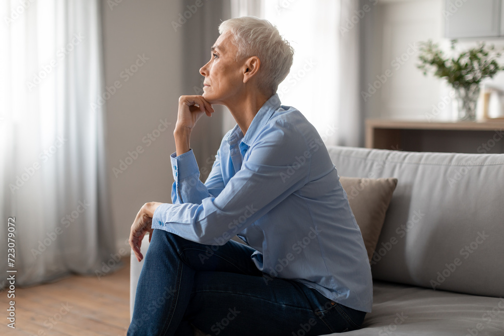 Depressed senior woman touching chin sitting on couch at home Depressed senior woman touching chin sitting on couch at home