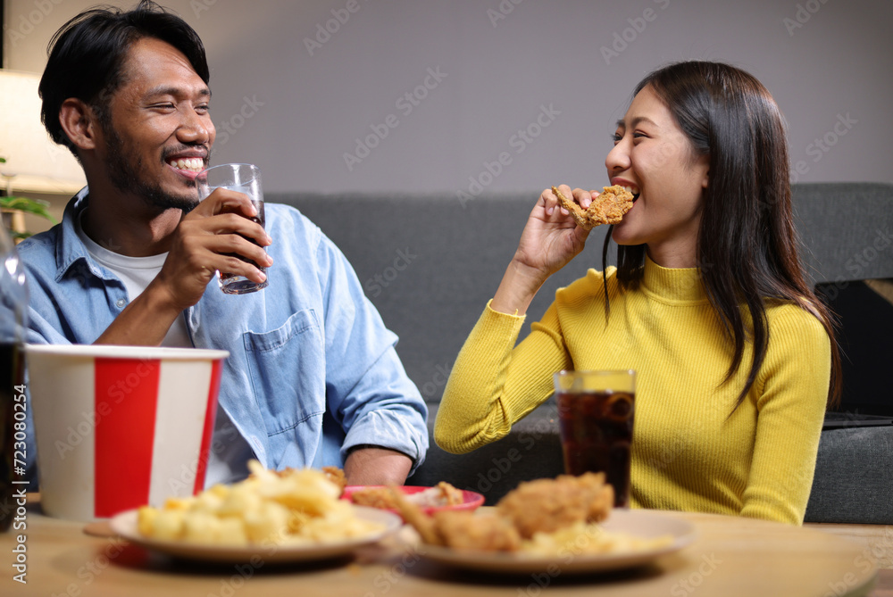 Young man and woman are eating dinner together at home. Friends eating fried chicken, drinks and snacks together while having dinner at home. Young man and woman are eating dinner together at home. Friends eating fried chicken, drinks and snacks together while having dinner at home.