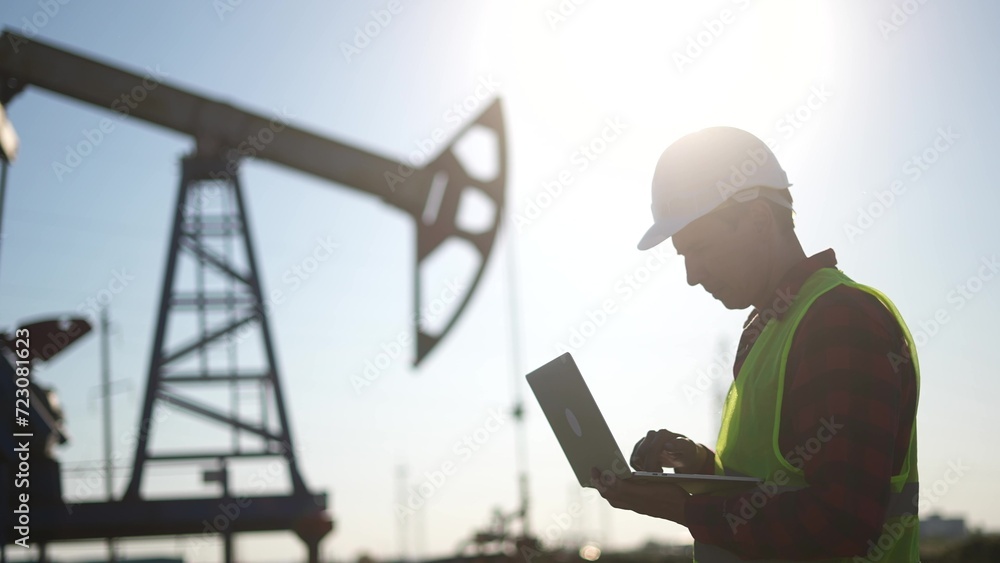 oil business. a worker works next to an oil pump holding a laptop. industry business lifestyle oil and gas concept. engineer studying the level of oil production on a laptop silhouette at sunset oil business. a worker works next to an oil pump holding a laptop. industry business lifestyle oil and gas concept. engineer studying the level of oil production on a laptop silhouette at sunset