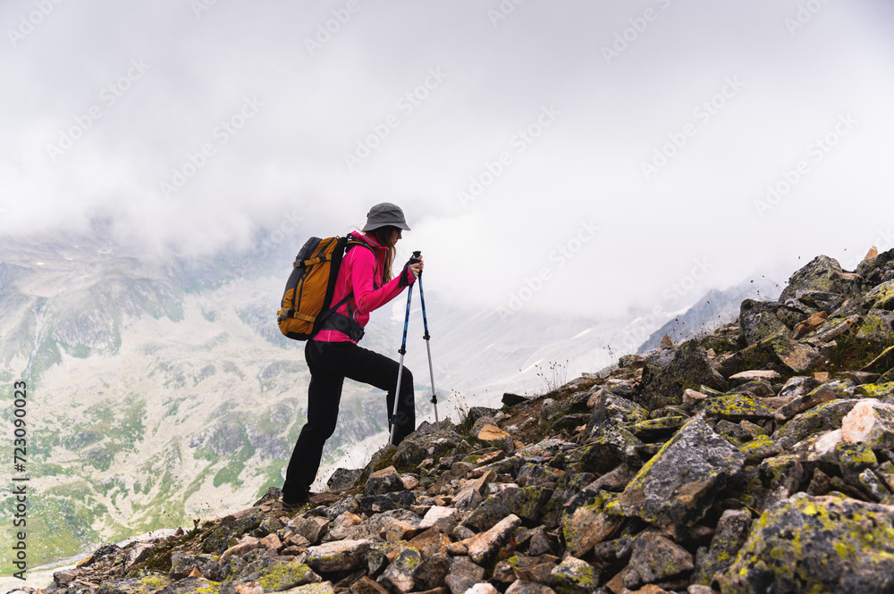 woman traveler goes to the top of the mountain. A girl with trekking poles and a backpack stopped to admire the fog approaching her in the mountains